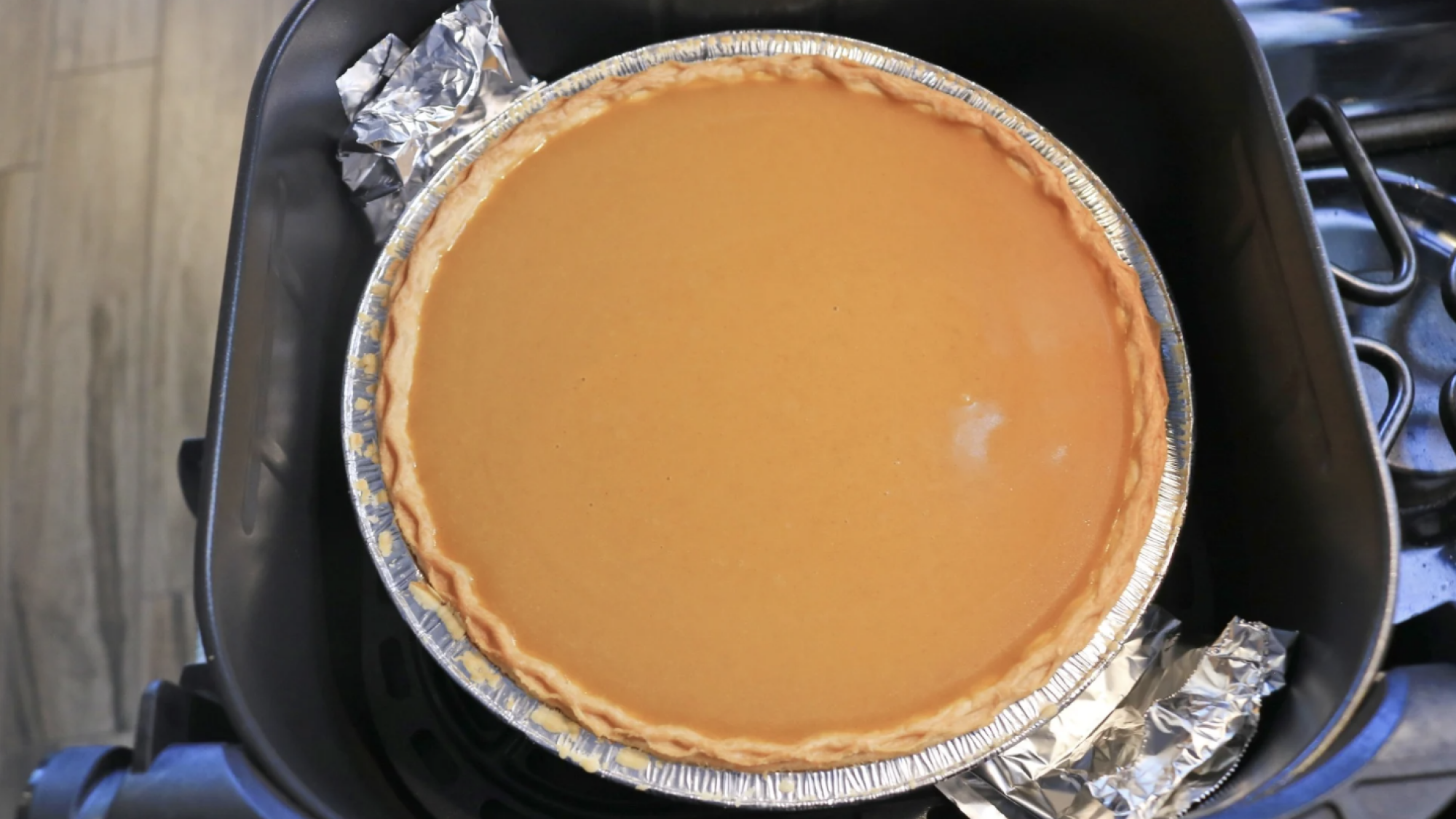 A pumpkin pie in an air fryer basket with a foil strip peeking out the sides.